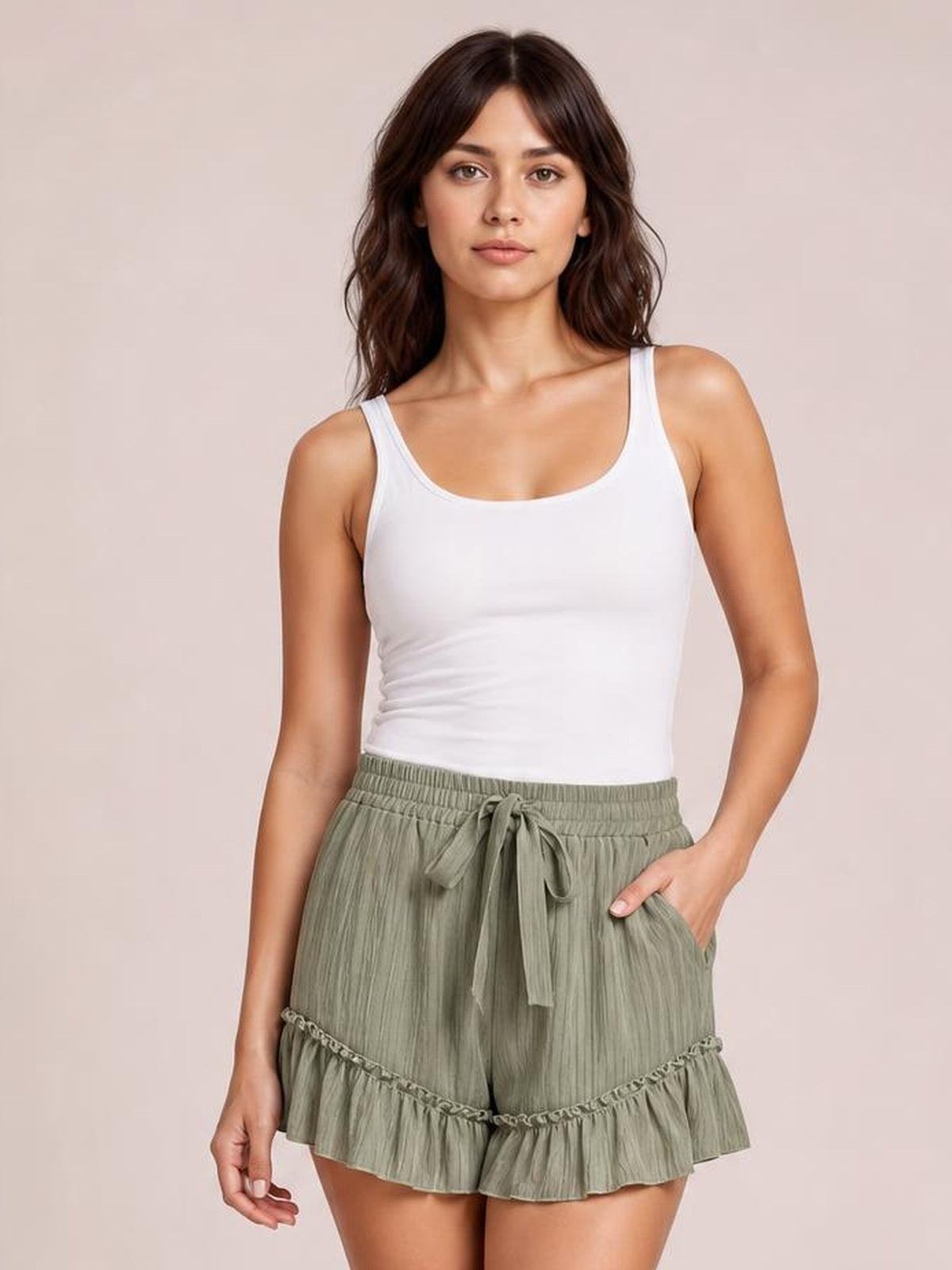 A woman modeling a white ribbed tank top and sage green pleated ruffled shorts with a drawstring waist, set against a solid light beige background.
