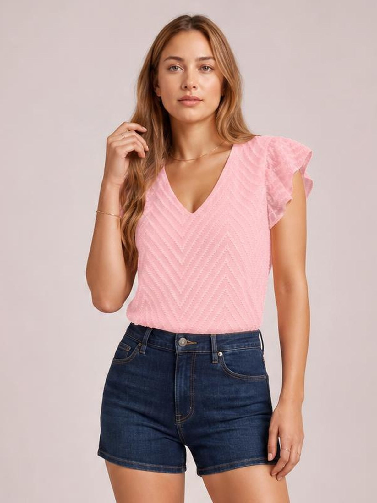 Model poses in a pink textured V-neck blouse with cap sleeves, a relaxed silhouette, and subtle chevron construction, tucked into dark denim shorts.