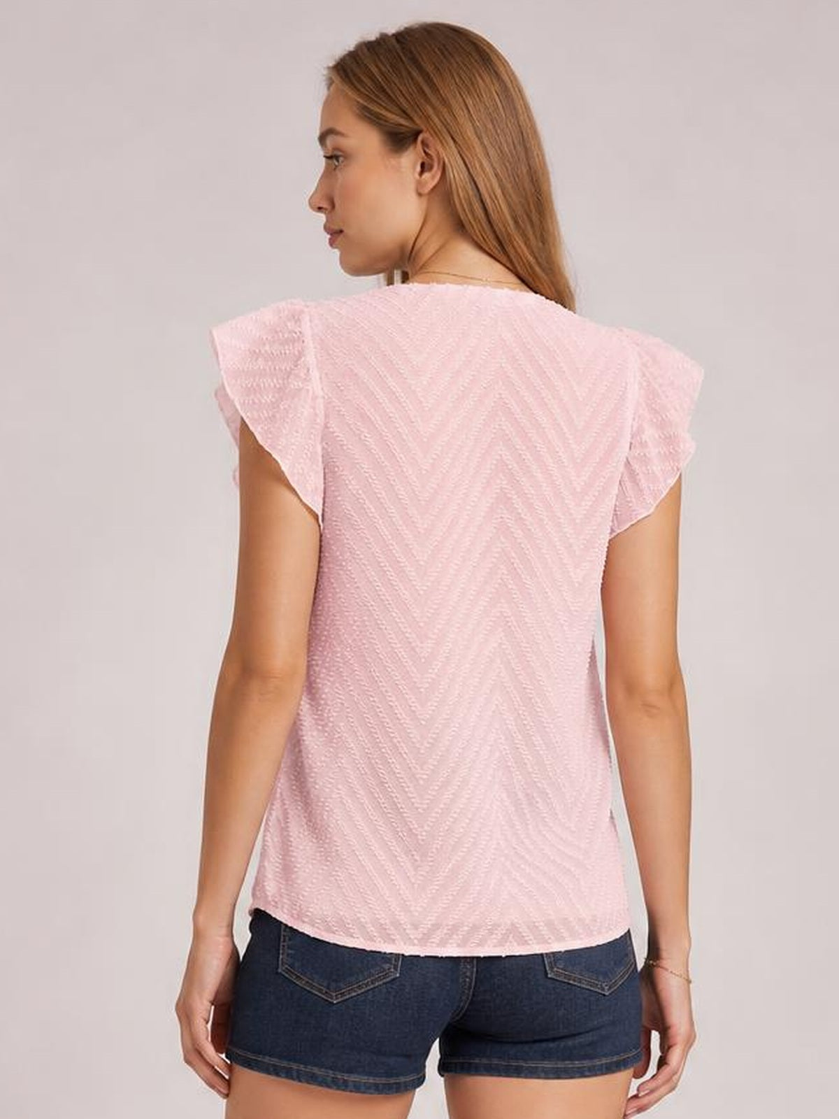 A model stands in a pale pink textured blouse with fluttering cap sleeves and a relaxed straight silhouette, shown from the back.