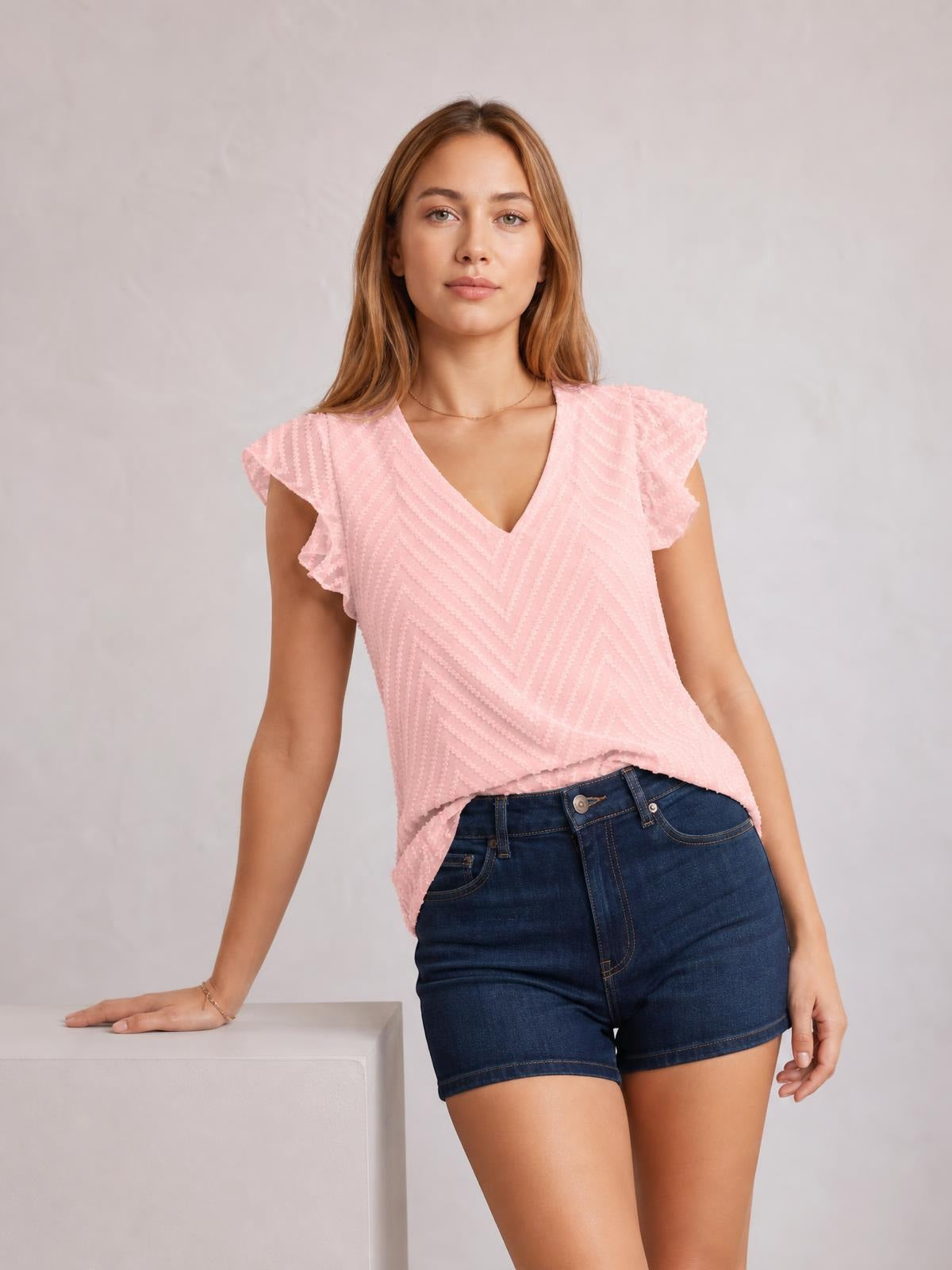 Model wearing a pink textured V-neck cap sleeve blouse with fluttered sleeve edges, a relaxed silhouette, and a slightly curved hem over denim shorts.