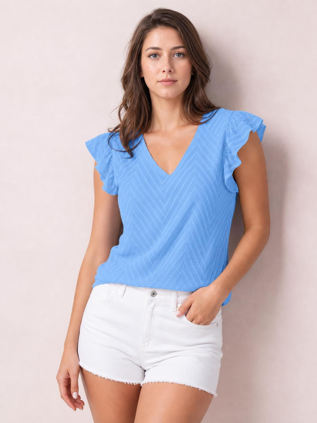 Model wears a light blue textured V-neck cap sleeve blouse with ruffled sleeves and a relaxed silhouette, posing with one hand in her white shorts pocket.