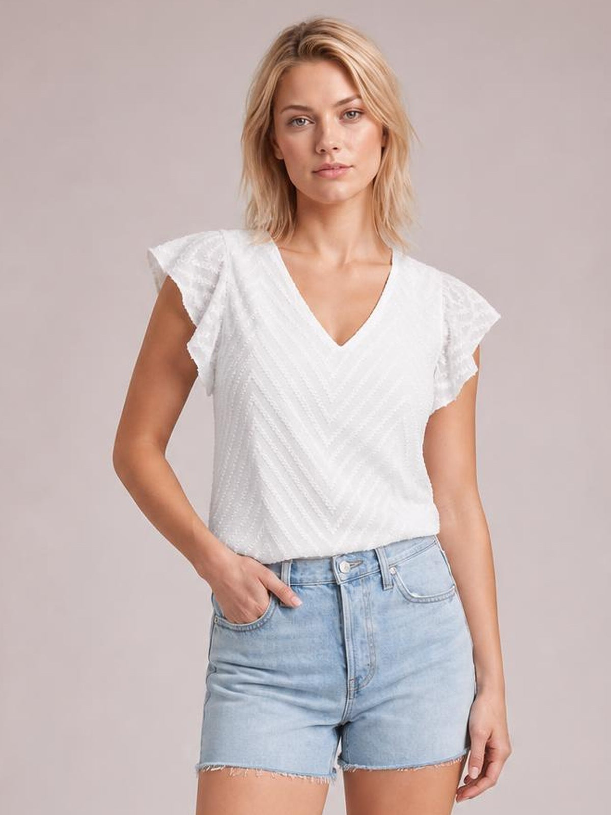 Model poses with one hand in her pocket wearing a white textured V-neck blouse with cap sleeves and a relaxed straight silhouette.