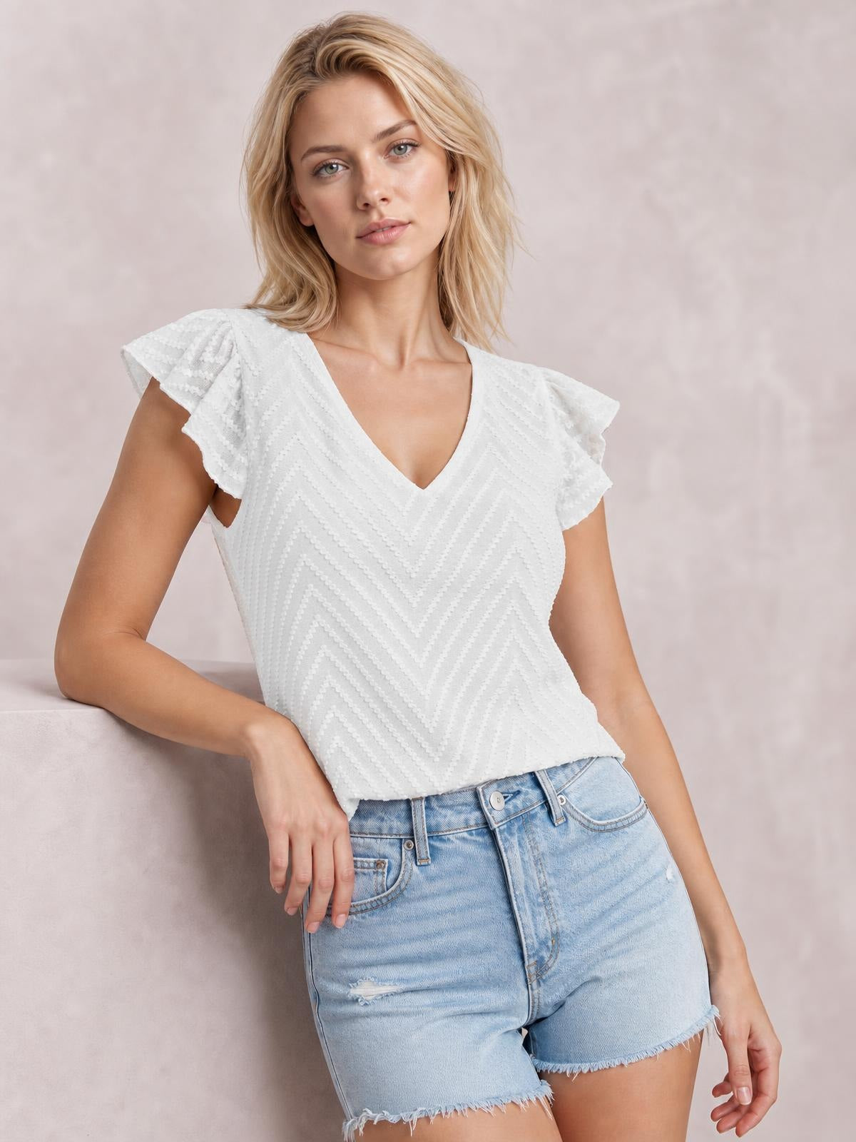 A model leans with one hand on a ledge wearing a white textured V-neck blouse with fluttering cap sleeves and a fitted silhouette.