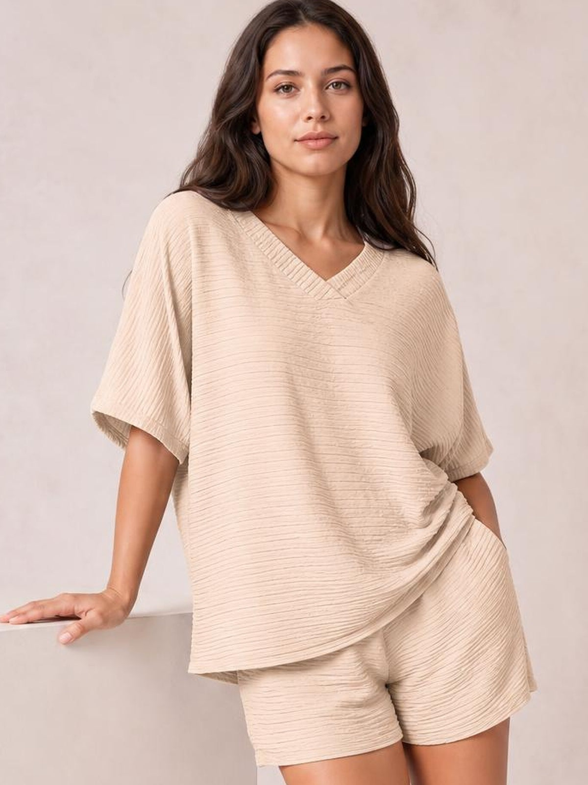 Model poses in a beige textured V-neck T-shirt and matching relaxed shorts set, featuring an oversized silhouette, short sleeves, and ribbed neckline.