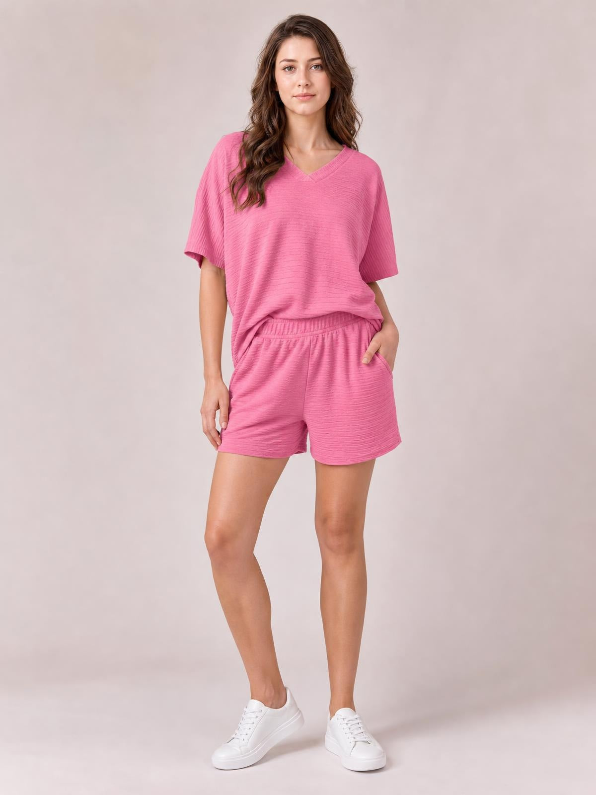 A woman poses in a relaxed pink textured V-neck T-shirt and shorts set with short sleeves, a loose silhouette, elastic waistband, and side pockets.