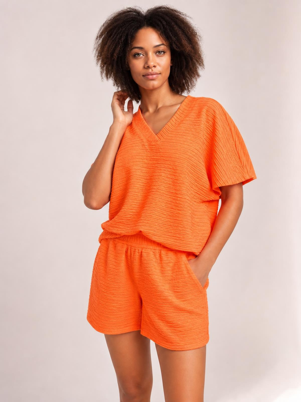 A model poses in a bright orange textured V-neck T-shirt and matching relaxed shorts, featuring short sleeves, an elastic waistband, and a side pocket.