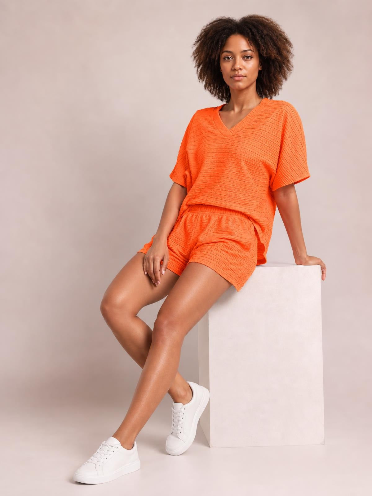 A model sits with crossed legs in a bright orange textured V-neck T-shirt and matching shorts set, featuring short sleeves and a relaxed silhouette.