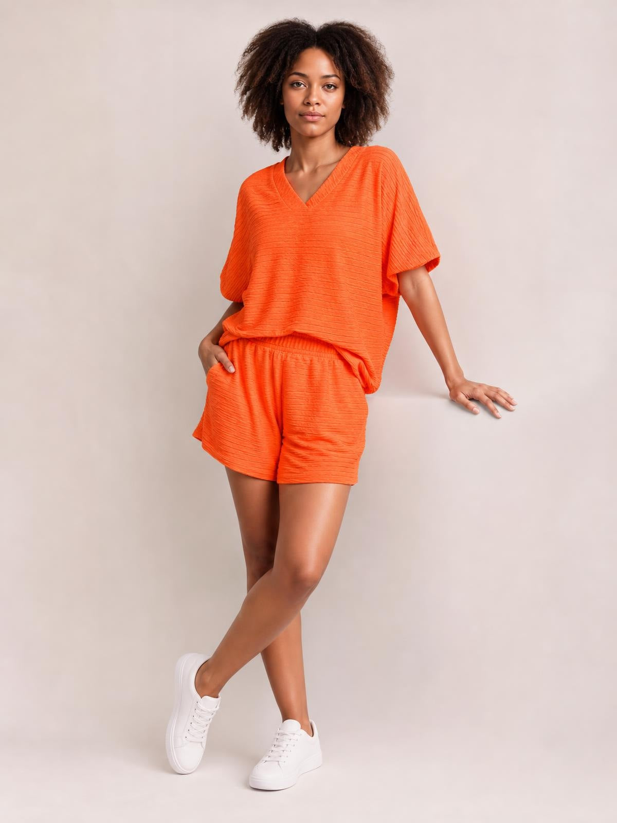 Model poses in an orange textured V-neck T-shirt and matching relaxed shorts set, with an easy silhouette, elastic waistband, and crossed legs.