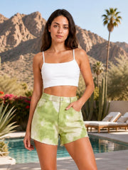 Model poses in green and white tie-dye high-rise shorts with belt loops, front button closure, pockets, and raw-cut hems.