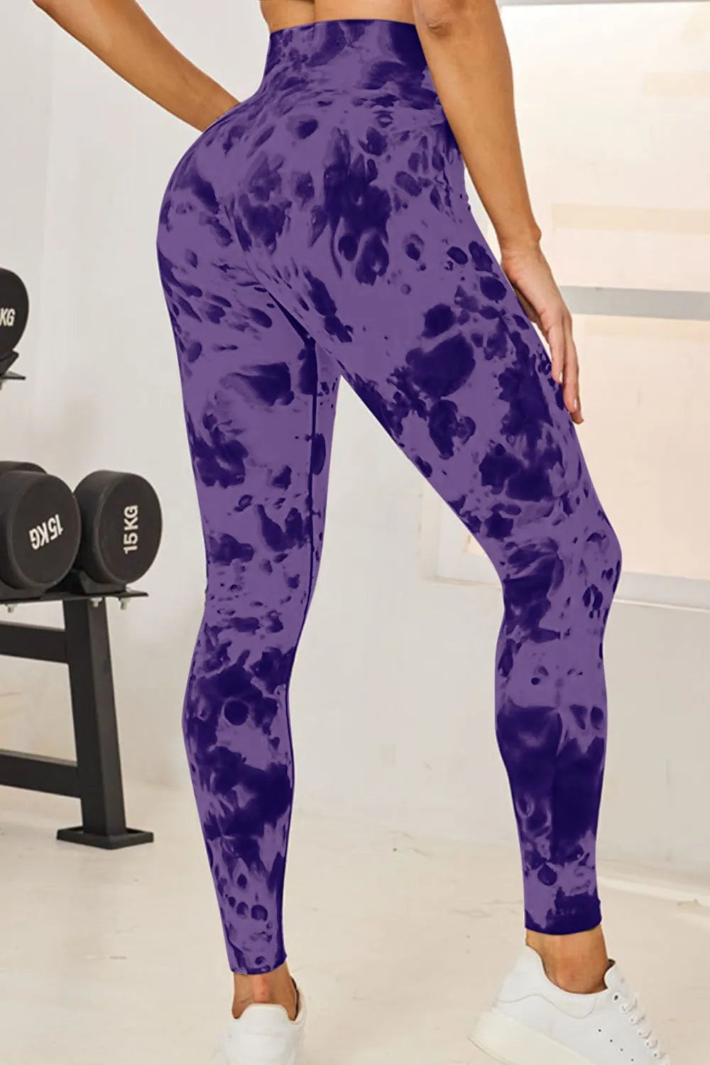 Model poses in purple tie-dye high-waist active leggings with a close, full-length silhouette and slightly tapered ankle fit.