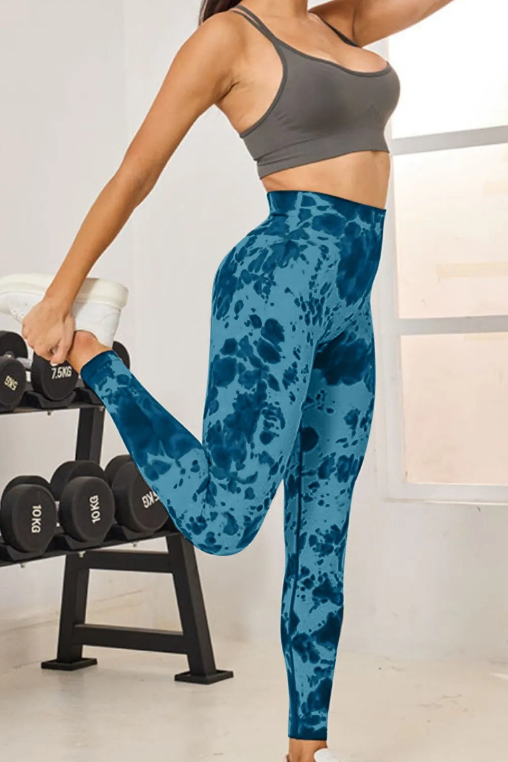 A model poses in blue tie-dye high-waist active leggings with a fitted, full-length silhouette, lifting one leg to show their stretch and sleek construction.
