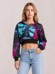 Model poses with one hand in her jeans pocket, wearing a cropped tie-dye hoodie in black, blue, and pink with a lace-up neckline and long sleeves.