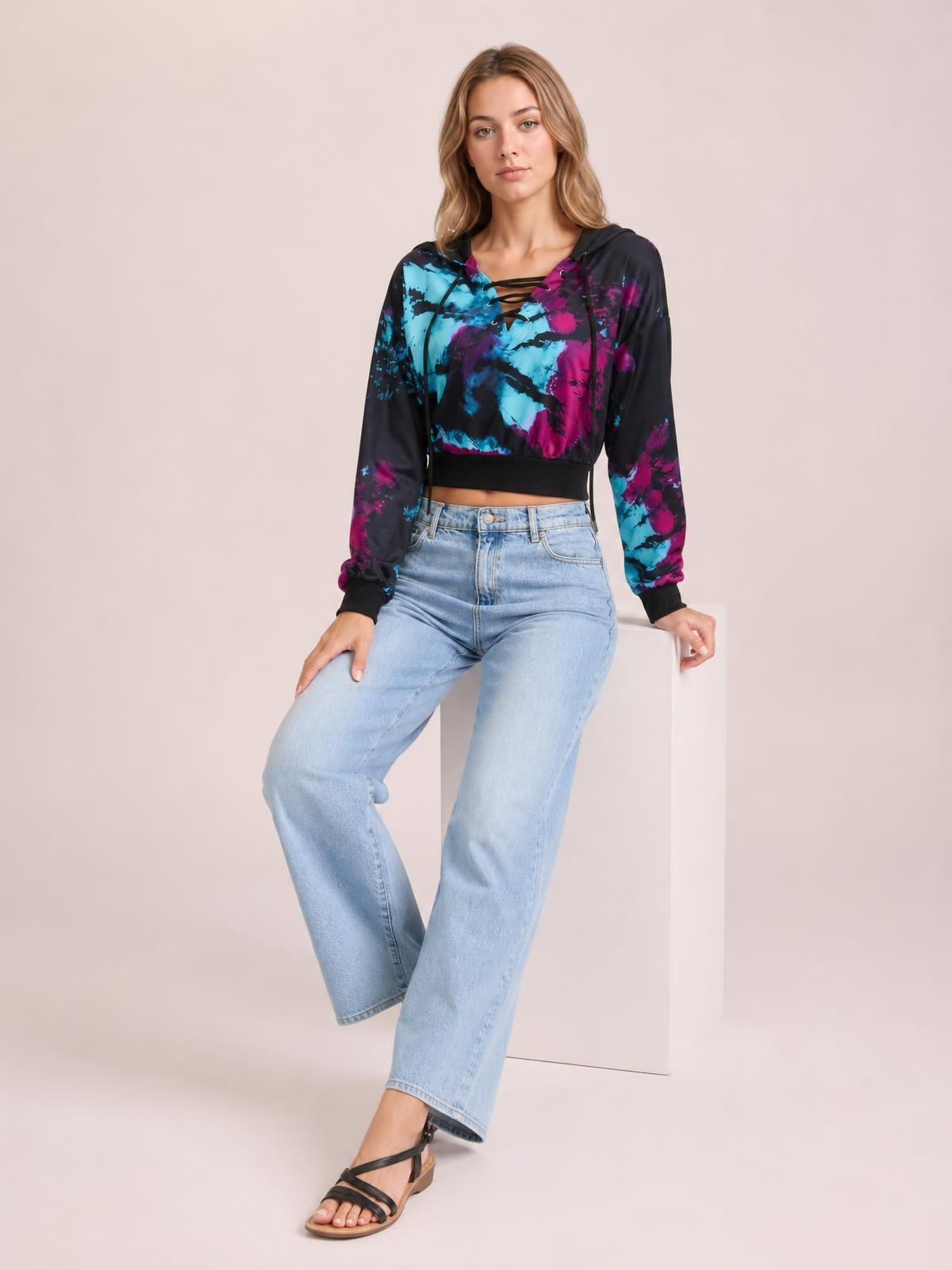 A model sits in a relaxed pose wearing a cropped tie-dye lace-up hoodie in black, blue, and magenta with long sleeves and a hood.