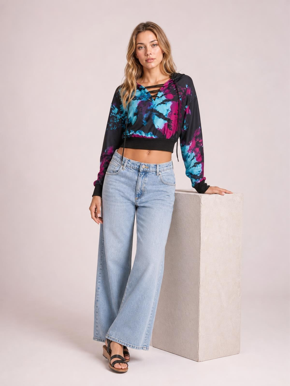 Model poses with one hand resting on a pedestal, wearing a cropped tie-dye lace-up hoodie in black, cyan, and magenta with long sleeves and ribbed trim.