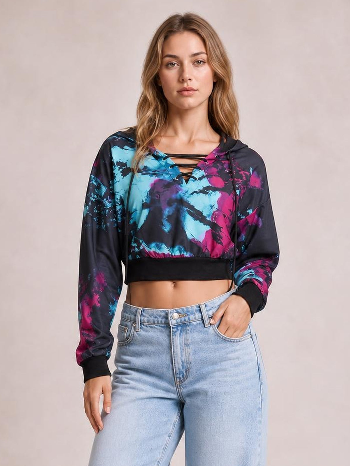 Model stands in a cropped tie-dye lace-up hoodie with a black base, vibrant blue and pink print, hood, long sleeves, and ribbed cuffs and hem.