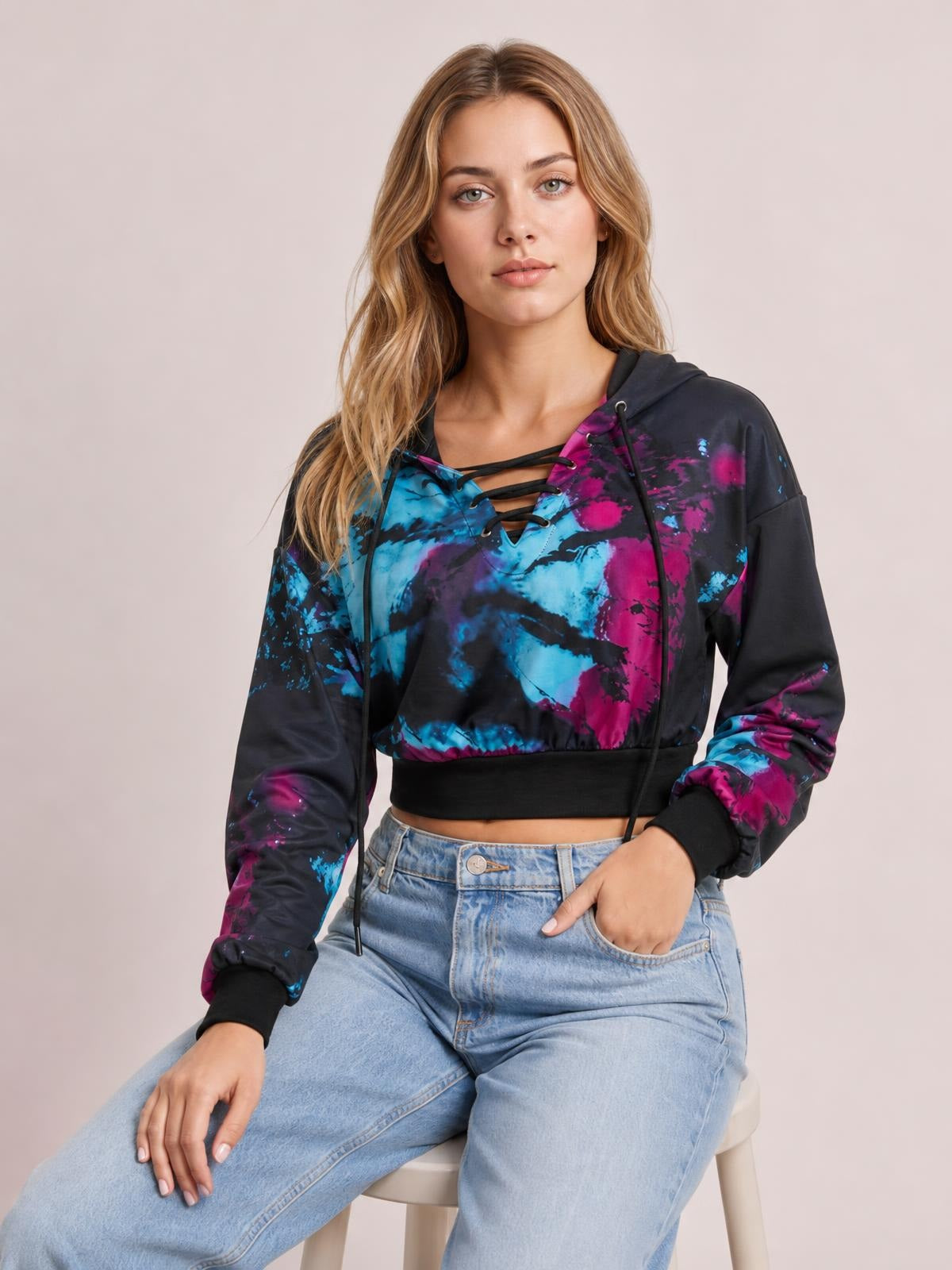 Seated model wears a black, blue, and magenta tie-dye lace-up hoodie with a drawstring hood, long sleeves, and a cropped silhouette.