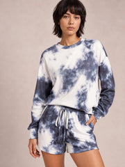 Model wearing a blue-and-white tie-dye lounge set with a round-neck long-sleeve top, dropped shoulders, and relaxed drawstring shorts, one hand in a pocket.