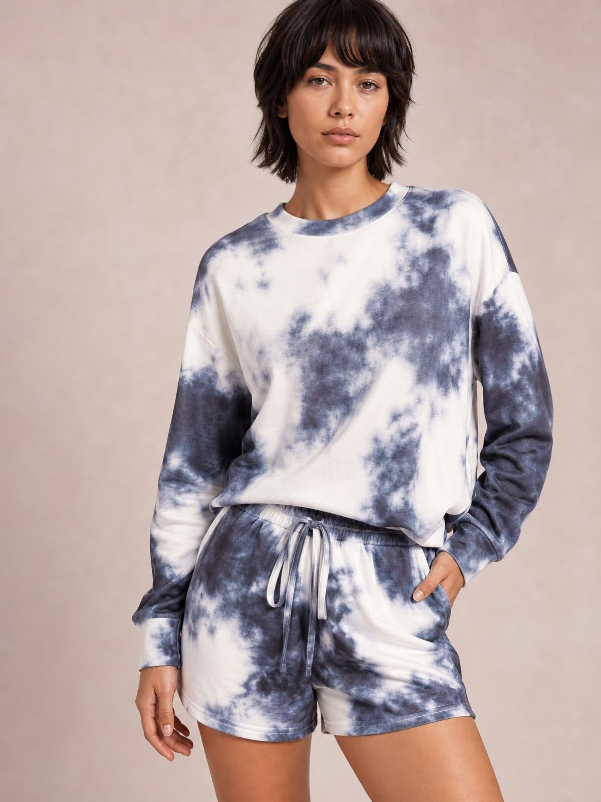Model wearing a blue-and-white tie-dye lounge set with a round-neck long-sleeve top, dropped shoulders, and relaxed drawstring shorts, one hand in a pocket.