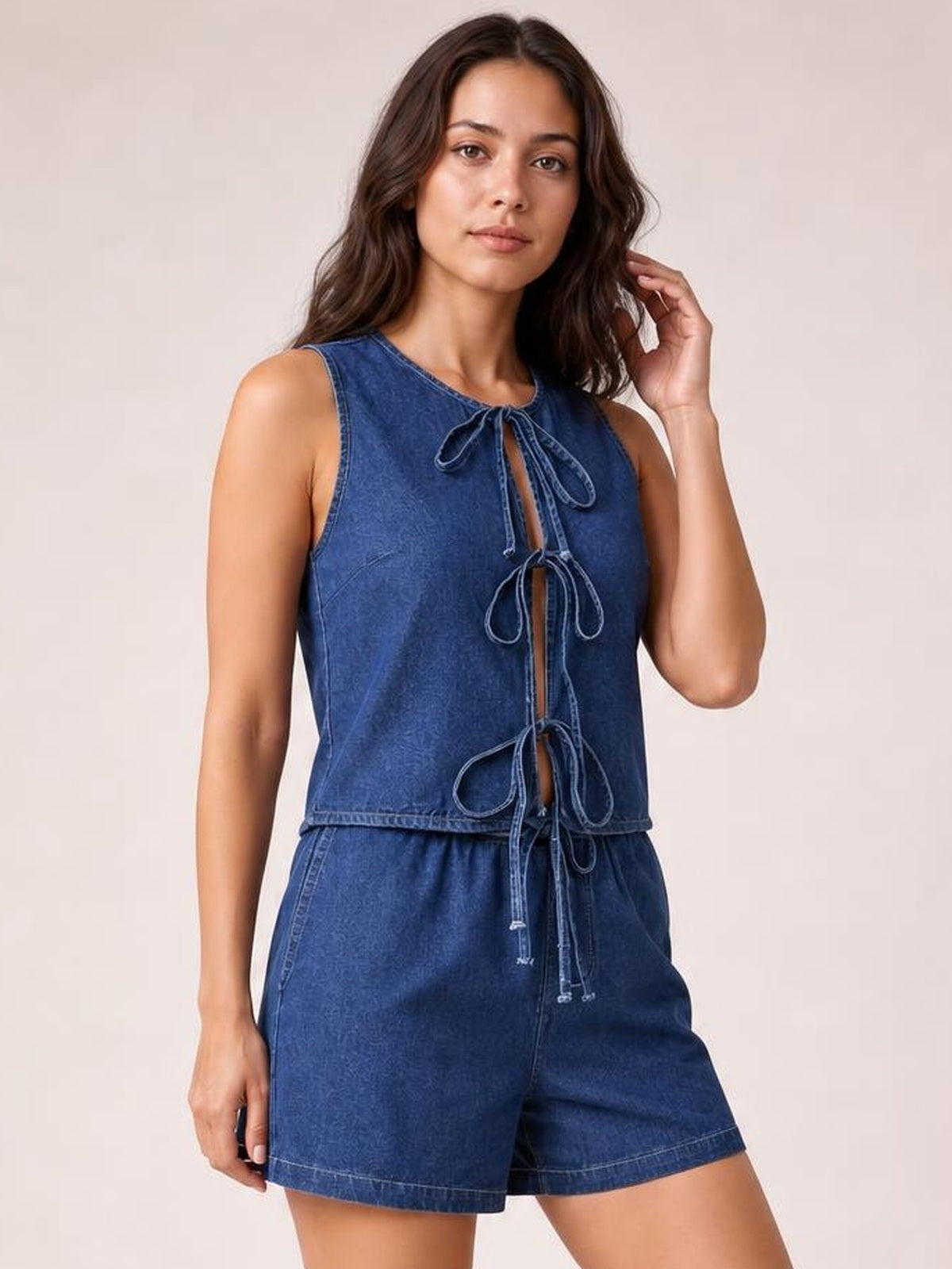 Model wears a blue denim set with a sleeveless tie-front top and relaxed shorts, posing with one hand touching her hair.