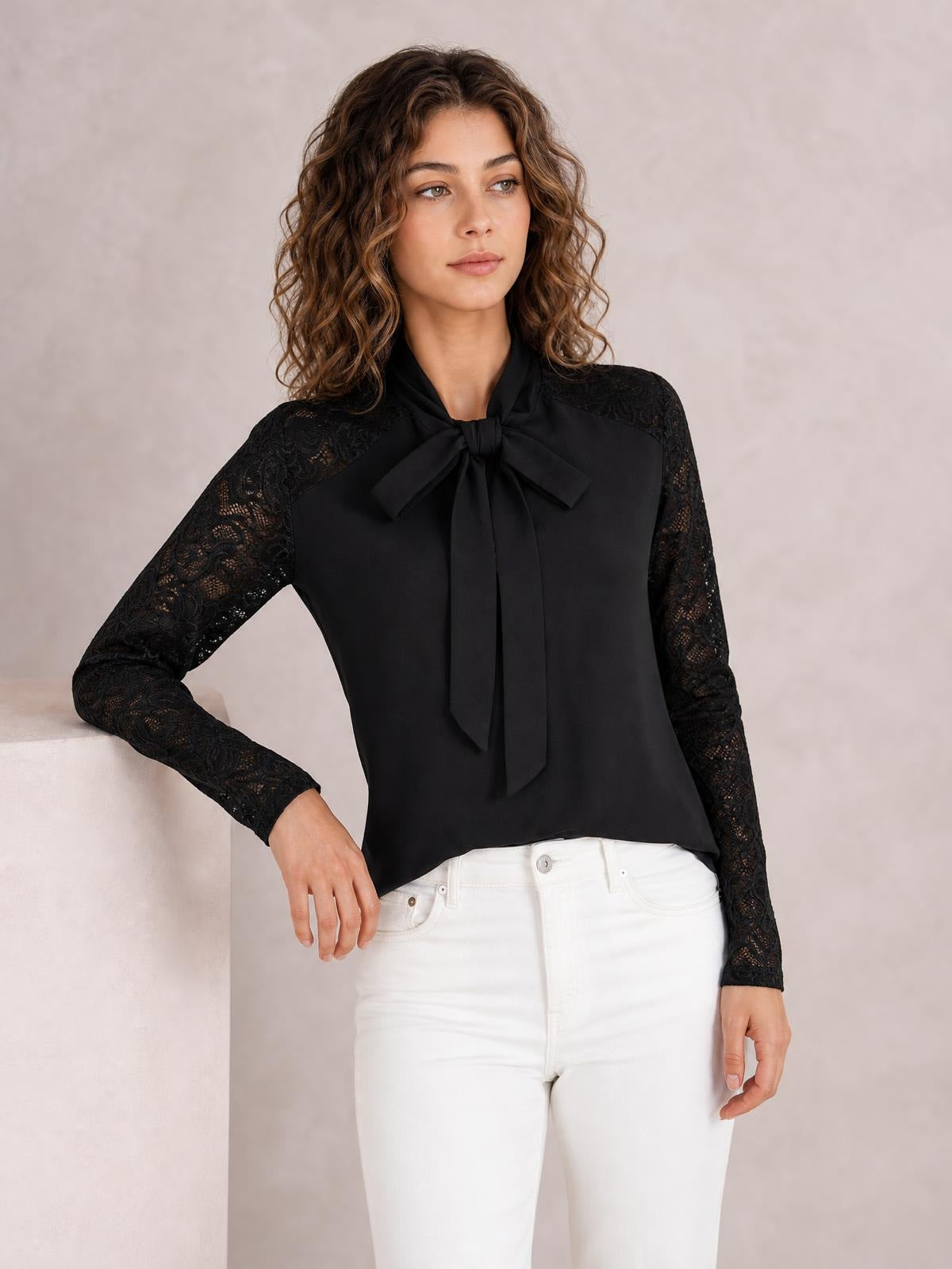 Model poses in a black tie-neck blouse with a bow front, solid smooth bodice, and contrasting floral lace long sleeves in a regular silhouette.