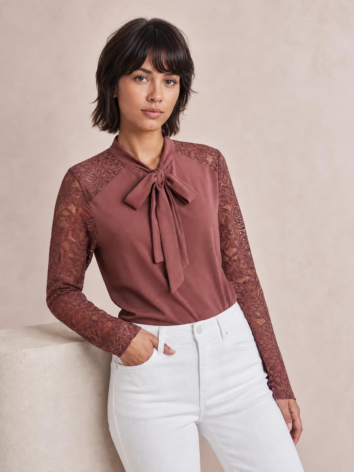 Model poses with one hand in her pocket wearing a solid mauve tie-neck top with a bow front, lace shoulder panels, long sleeves, and a regular silhouette.