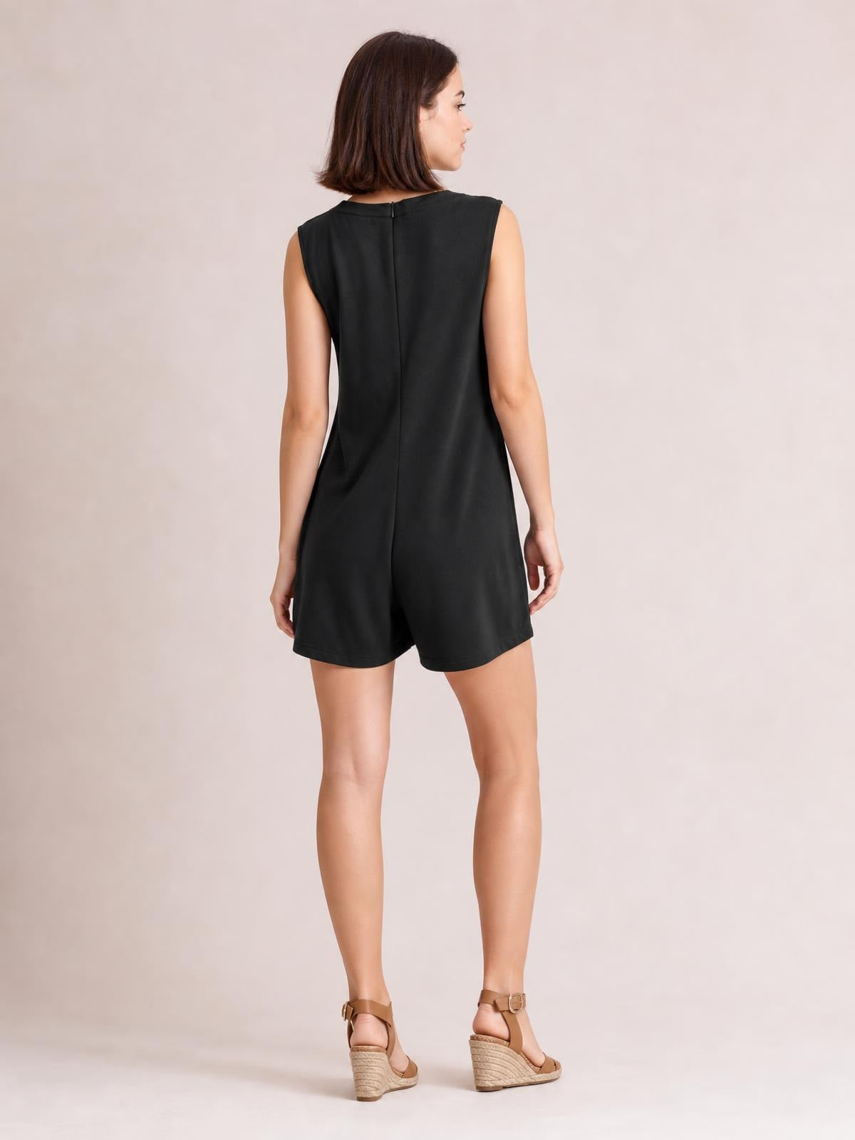 A model stands in a black sleeveless loose romper with a relaxed silhouette, shorts-length legs, and a visible center-back zipper.