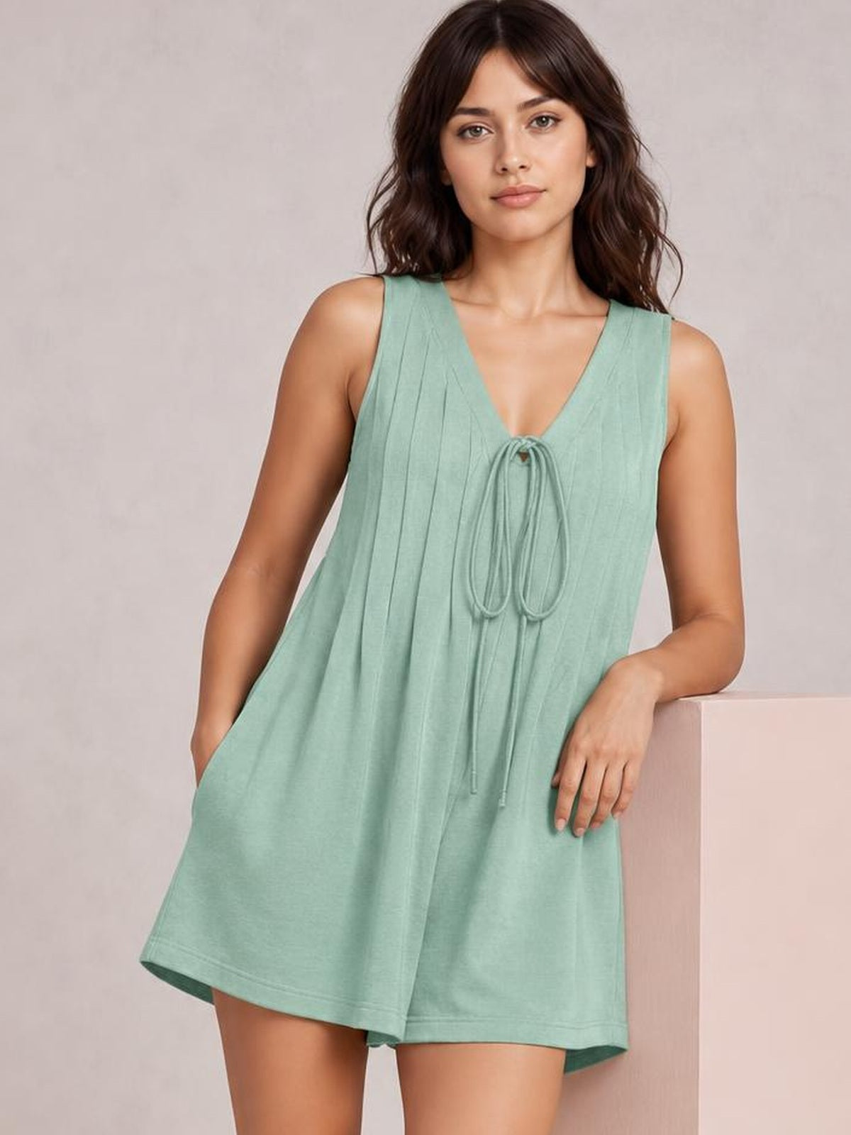 Model wears a mint green sleeveless loose romper with a tie V-neck, vertical pintuck detailing, and a relaxed wide-leg silhouette.