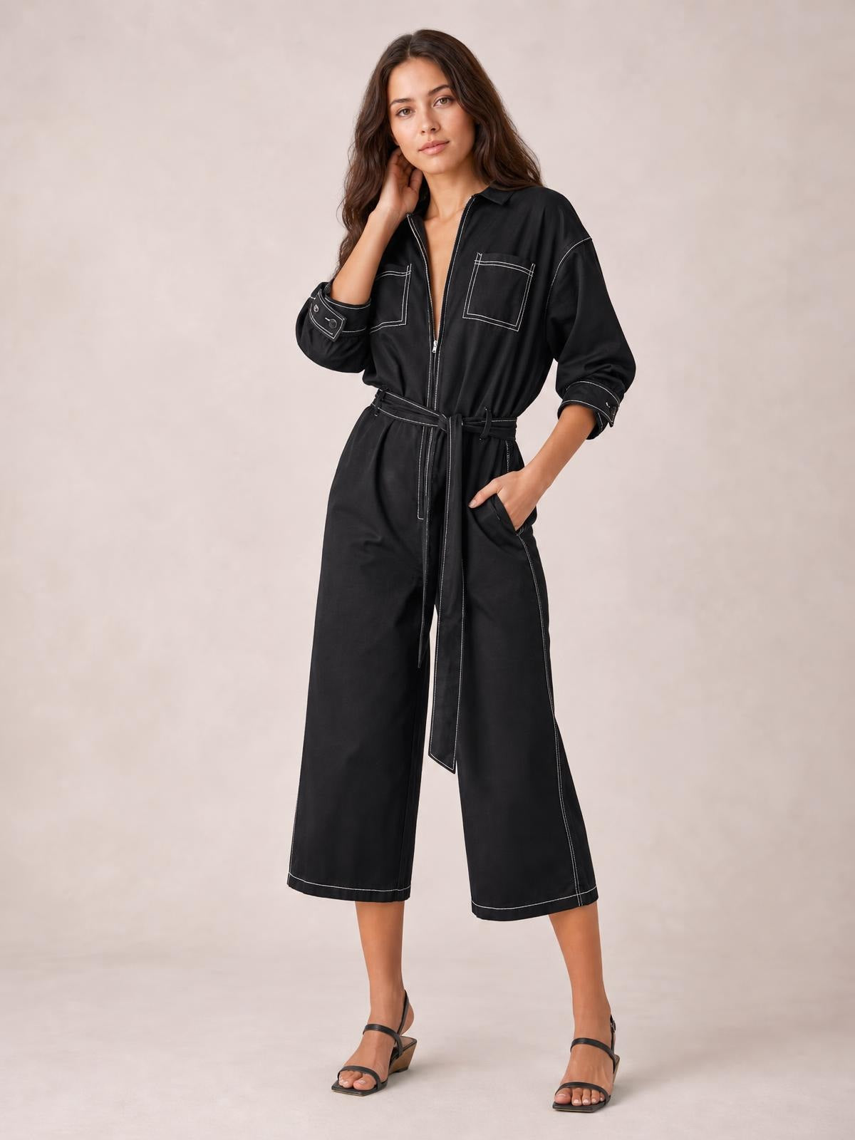 Model poses in a black tie waist wide leg jumpsuit with white contrast stitching, zip front, chest pockets, rolled cuffs, and cropped wide-leg trousers.