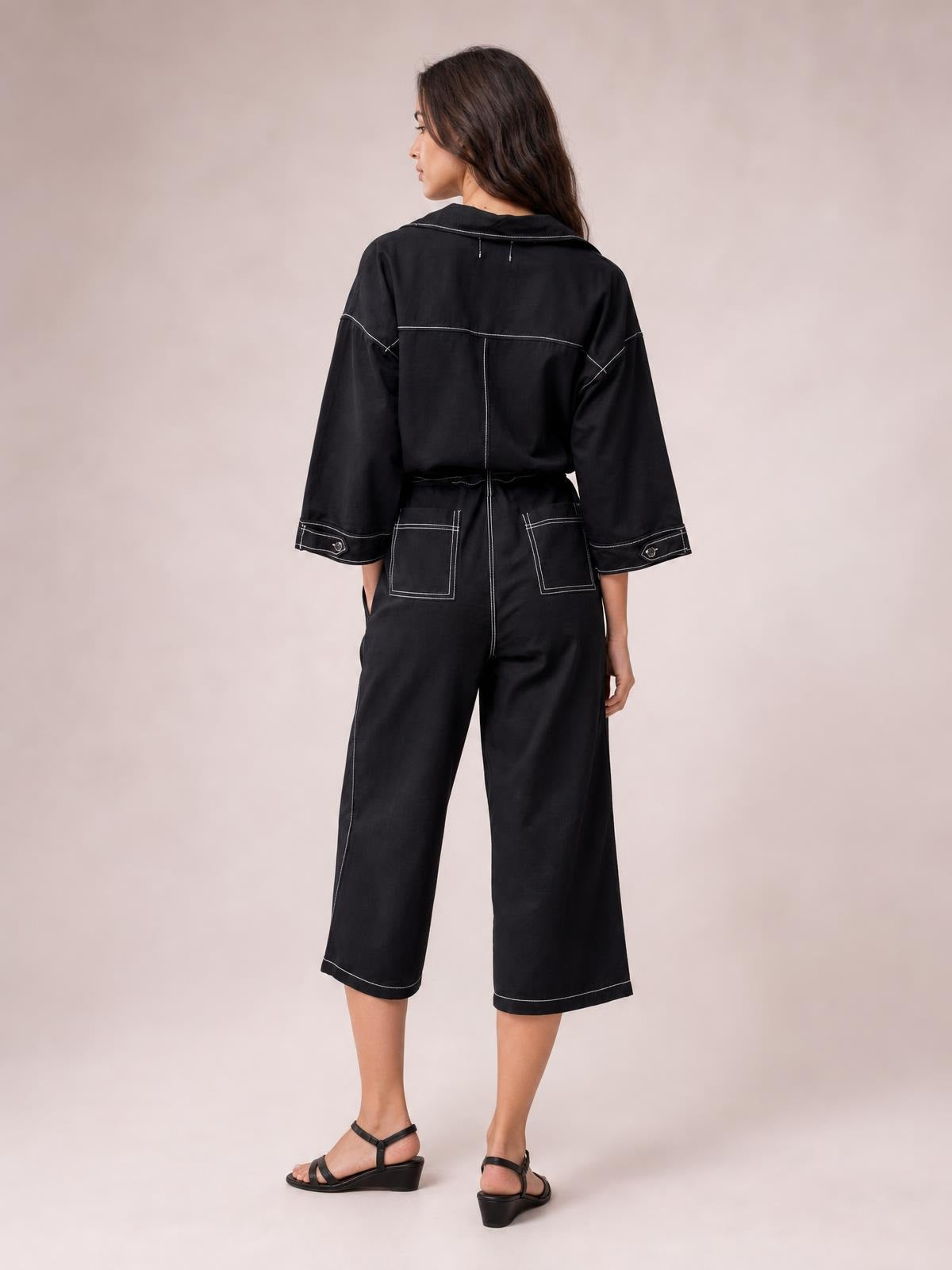 A model poses turned away in a black tie-waist jumpsuit with wide-leg cropped pants, long sleeves, contrast stitching, and back patch pockets.