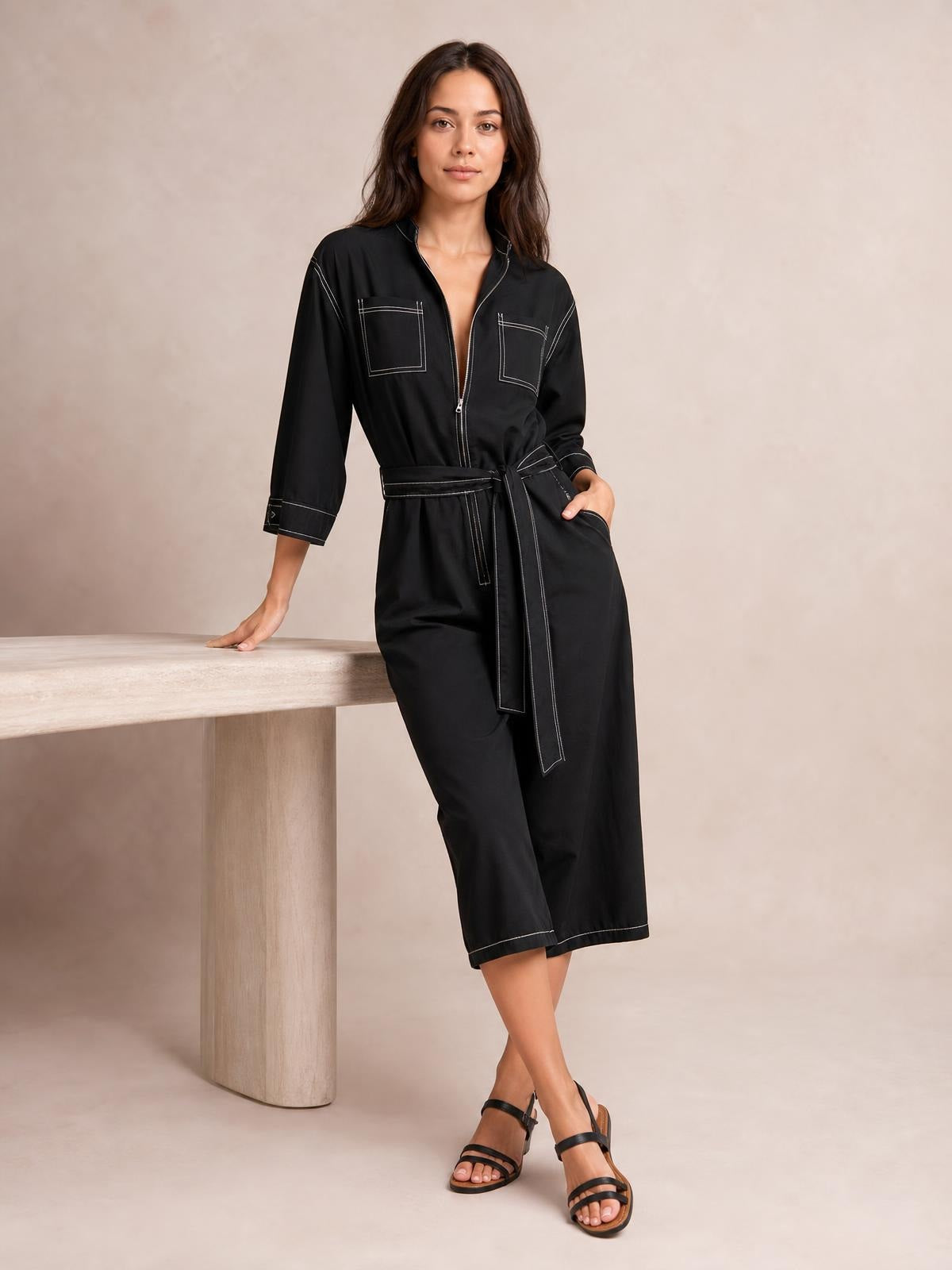 Model poses in a black tie waist wide leg jumpsuit with cropped legs, front zipper, patch pockets, contrast stitching, and one hand in a pocket.