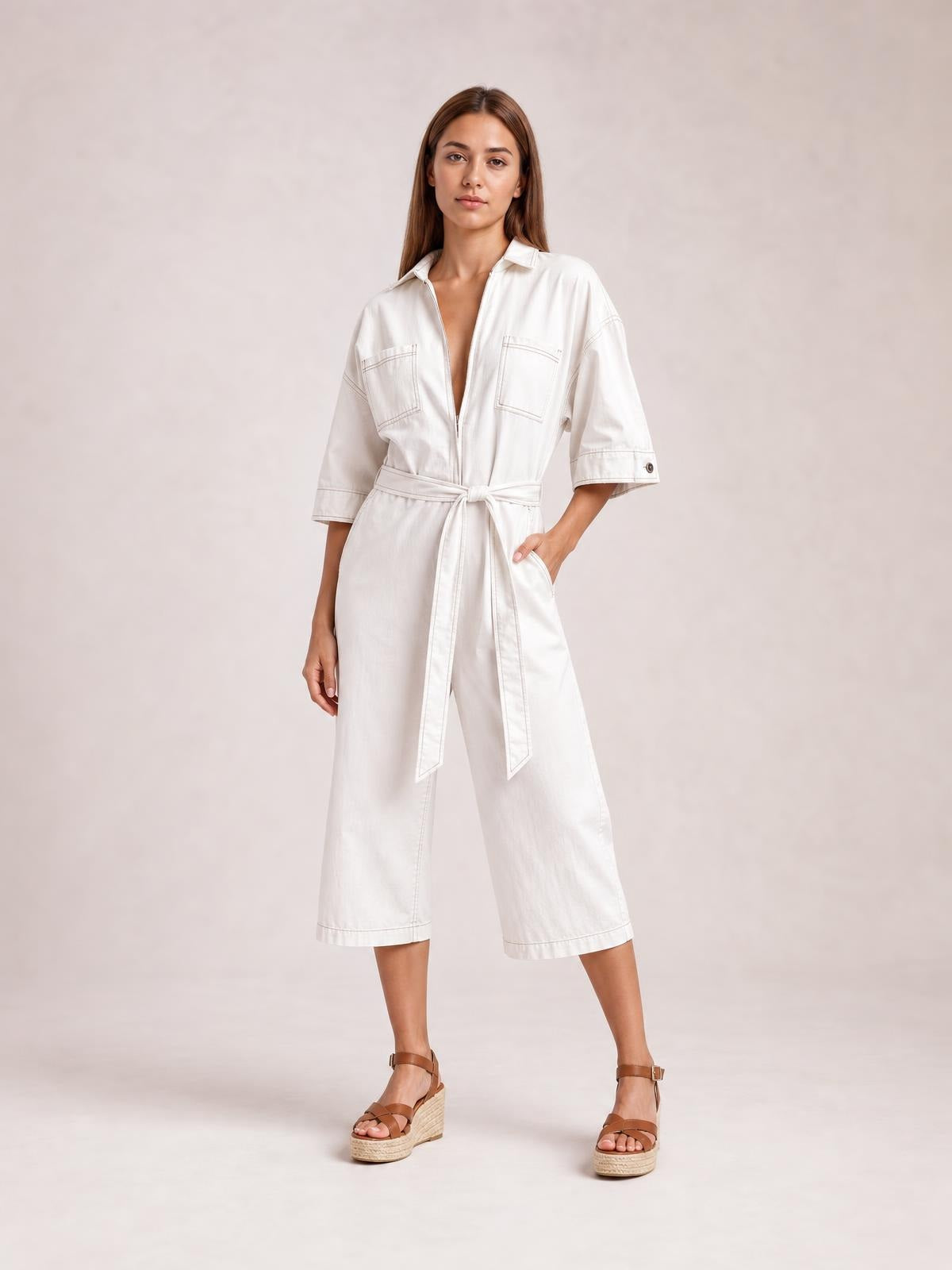 Model poses in a white tie-waist jumpsuit with a collared zip front, short sleeves, chest and side pockets, and cropped wide legs.