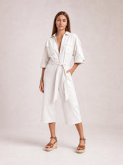 Model poses in a white tie-waist jumpsuit with a collared zip front, short sleeves, chest and side pockets, and cropped wide legs.