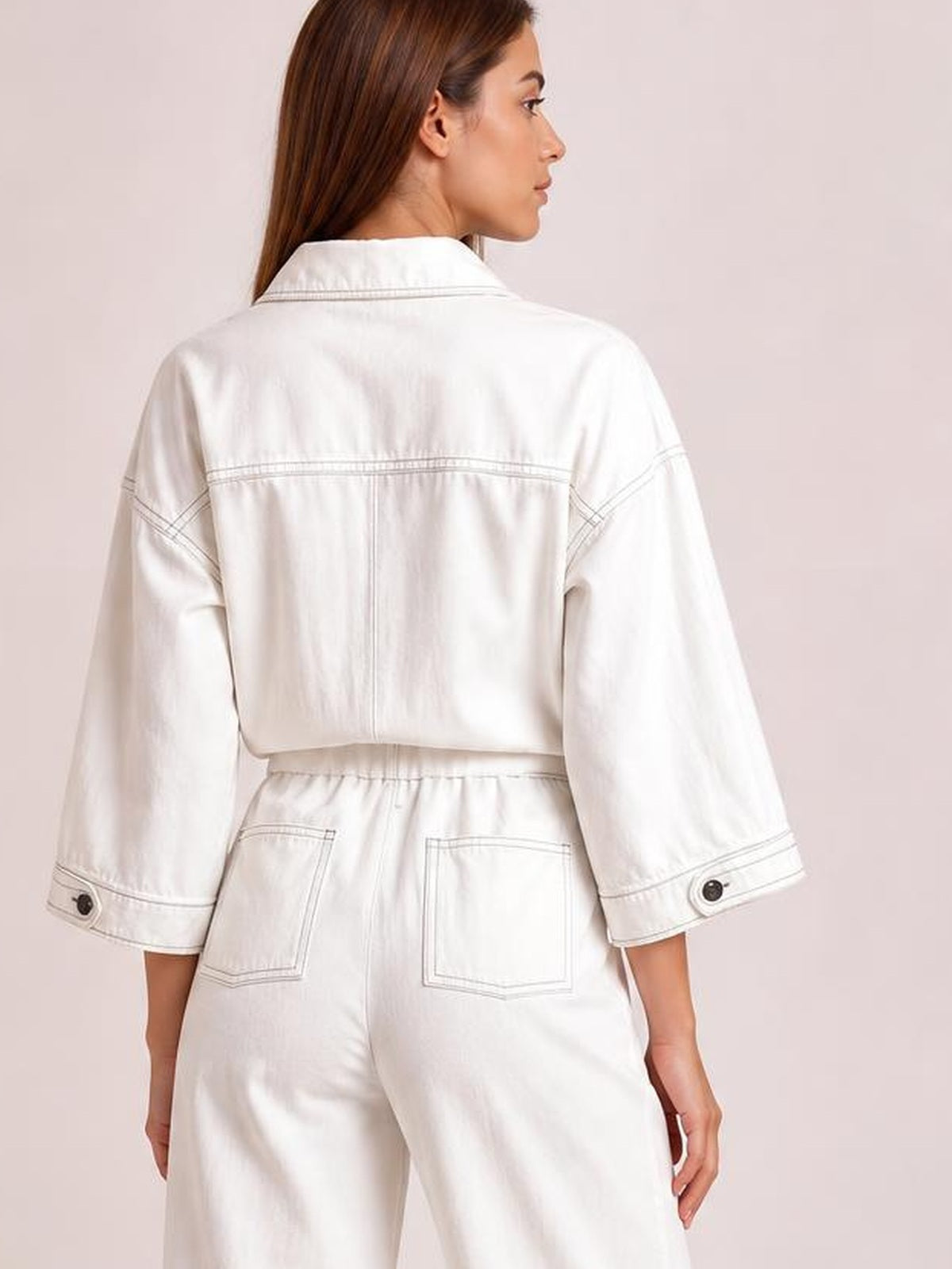 A model faces away wearing a white tie-waist wide-leg jumpsuit with a collared long-sleeve top, cuff buttons, contrast stitching, and back pockets.