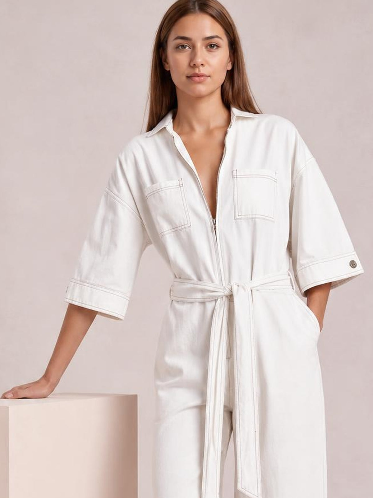 A model poses in a white tie-waist wide-leg jumpsuit with a collared zip front, patch pockets, and relaxed three-quarter sleeves.