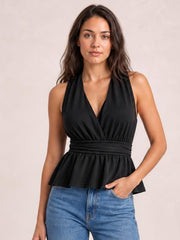 A model wears a black sleeveless peplum top with a deep V-neckline, wide gathered waistband, and flared hem, posing with one hand in her jeans pocket.