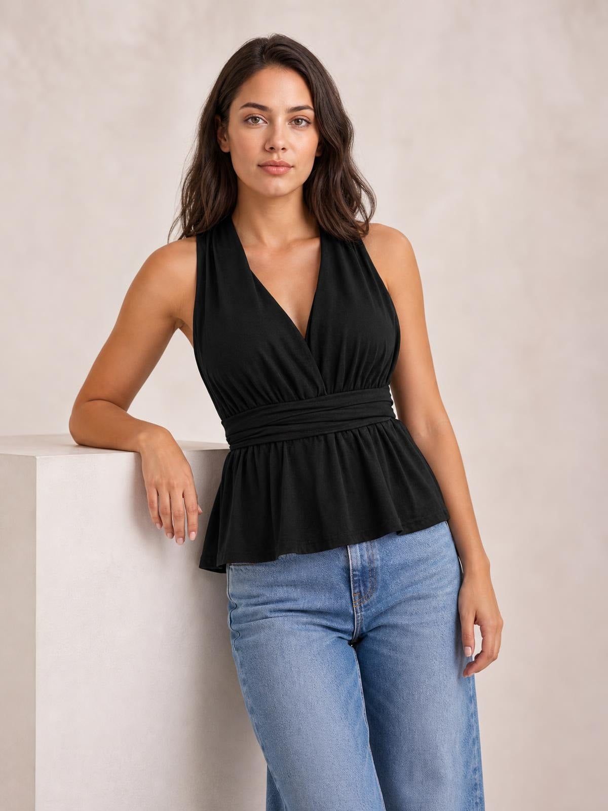 A model poses in a black sleeveless peplum top with a deep V-neck, gathered waist, and flared silhouette, paired with light-wash jeans.