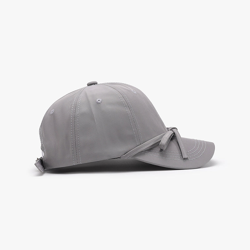 Gray cotton baseball cap with a tied bow across the front, curved brim, softly rounded paneled crown, neat topstitching, and an adjustable back strap.