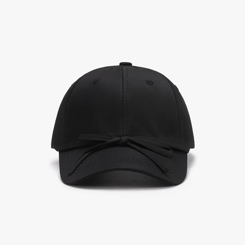 Black cotton baseball cap with a softly rounded paneled crown, curved brim, neat topstitching, and a tied bow detail across the front.