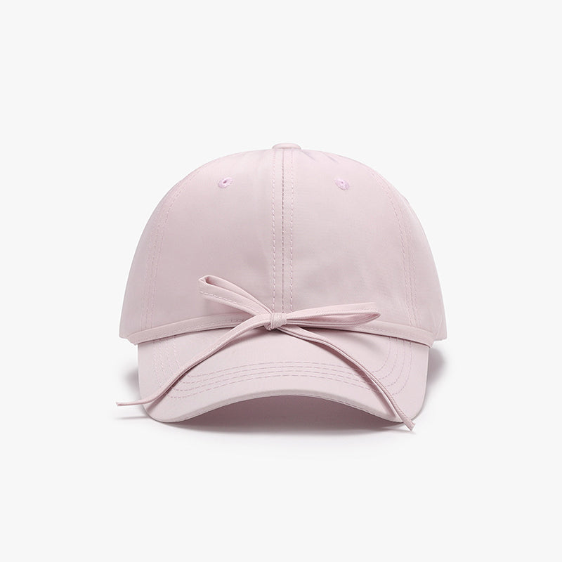 Light pink cotton baseball cap with a softly rounded paneled crown, curved brim, neat topstitching, and a tied bow detail across the front.