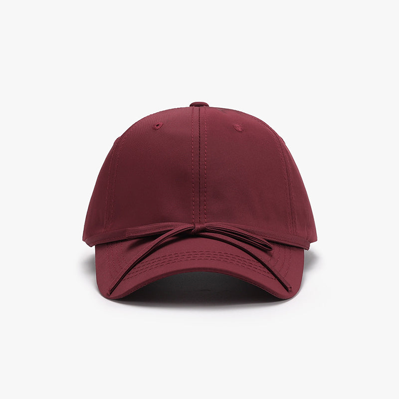 Burgundy cotton baseball cap with a softly rounded paneled crown, curved brim, neat topstitching, and a tied bow detail across the front.