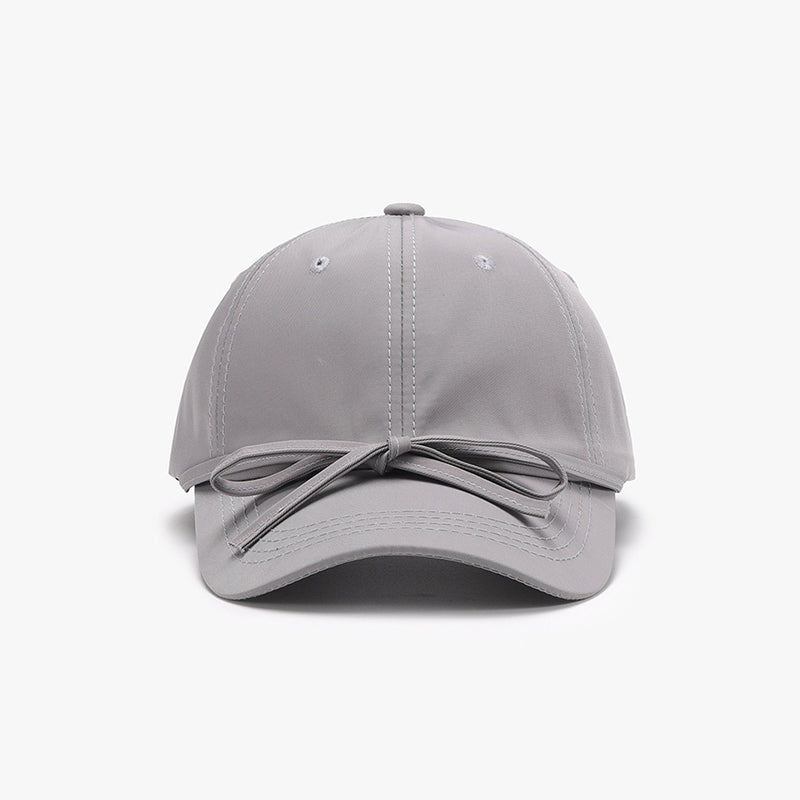 Gray cotton baseball cap with a softly rounded paneled crown, curved brim, neat topstitching, and a tied bow detail across the front.