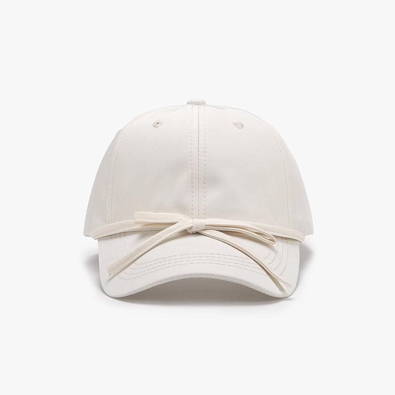 Ivory cotton baseball cap with a softly rounded paneled crown, curved brim, neat topstitching, and a feminine tied bow detail across the front.