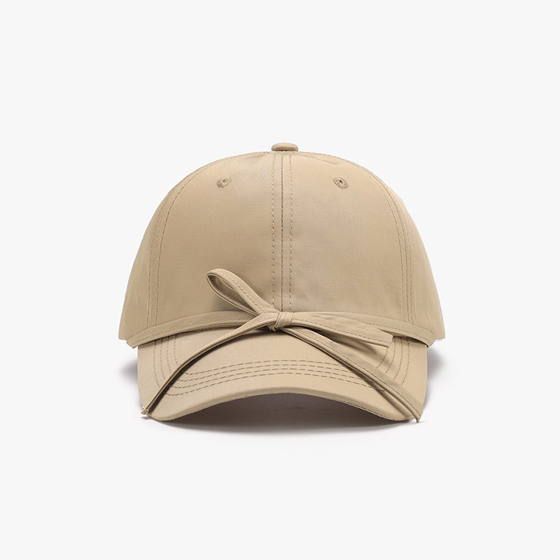 Beige cotton baseball cap shown front-on with a softly rounded paneled crown, curved brim, neat topstitching, and a tied bow detail across the front.