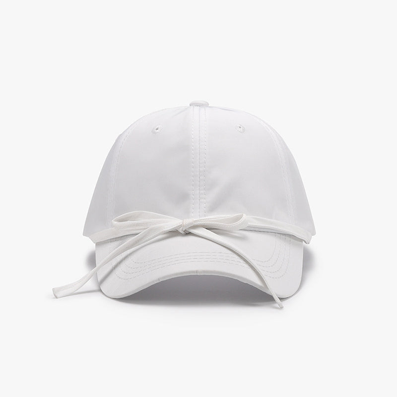 White cotton baseball cap with a softly rounded paneled crown, curved brim, neat topstitching, and a tied bow detail across the front.
