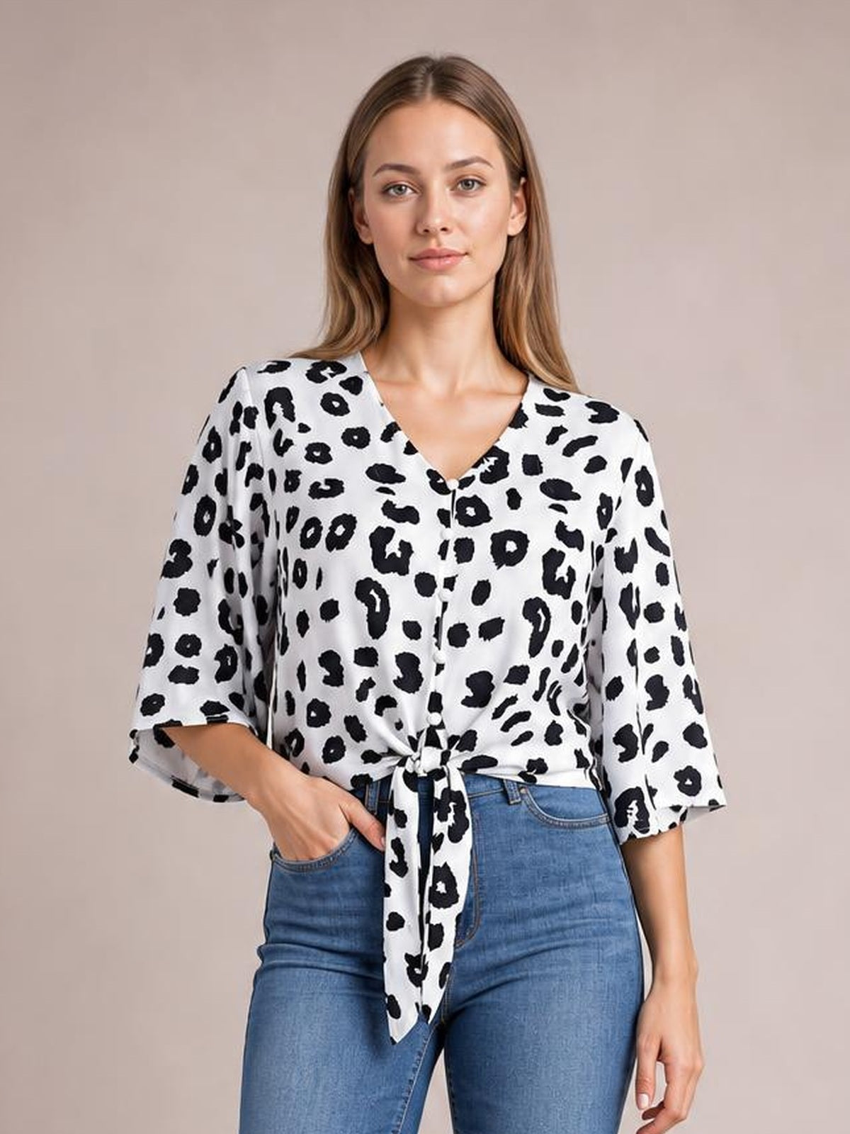 Model wearing a white and black leopard V-neck blouse with front buttons, tied waist detail, and loose three-quarter sleeves, posed with one hand in her jeans pocket.