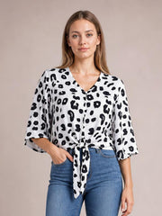 Model wearing a white and black leopard V-neck blouse with front buttons, tied waist detail, and loose three-quarter sleeves, posed with one hand in her jeans pocket.