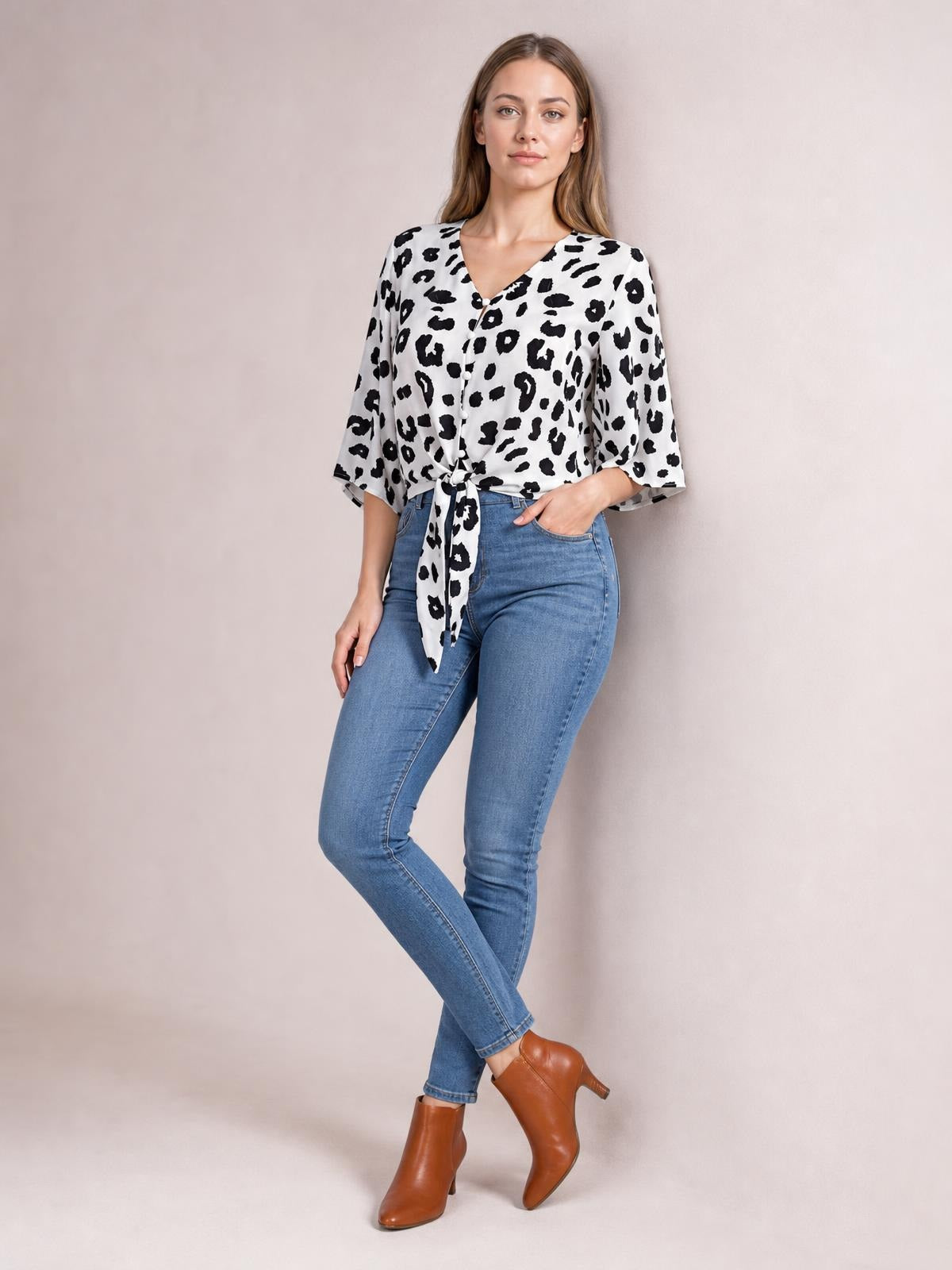 Model poses with one hand in her pocket, wearing a white and black leopard V-neck blouse with front buttons, a waist tie, and loose three-quarter sleeves.