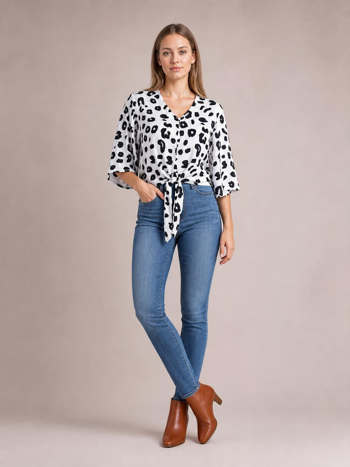Model poses with one hand in her jeans pocket, wearing a black-and-white leopard-print blouse with a V-neck, button front, three-quarter sleeves, and tied waist.