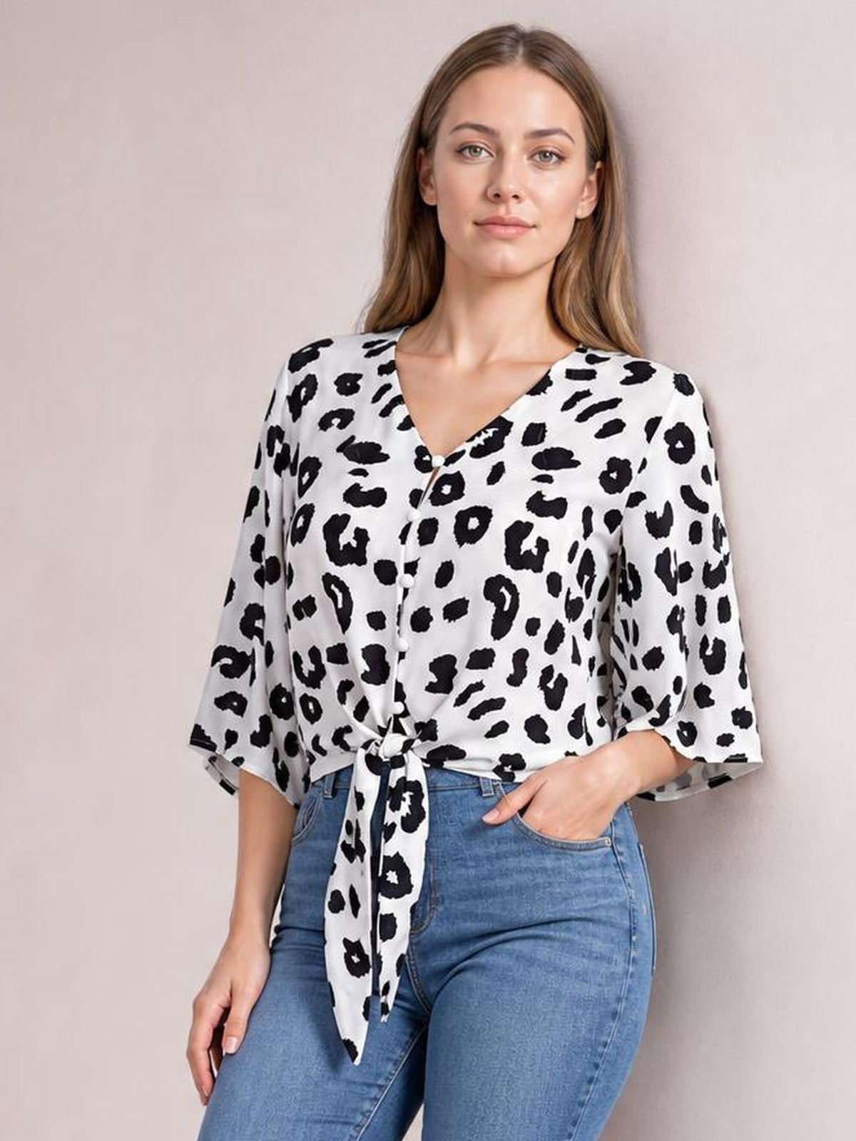 Model wears a black-and-white leopard V-neck blouse with button front, tied waist detail, and loose three-quarter sleeves, posing with one hand in her jeans pocket.