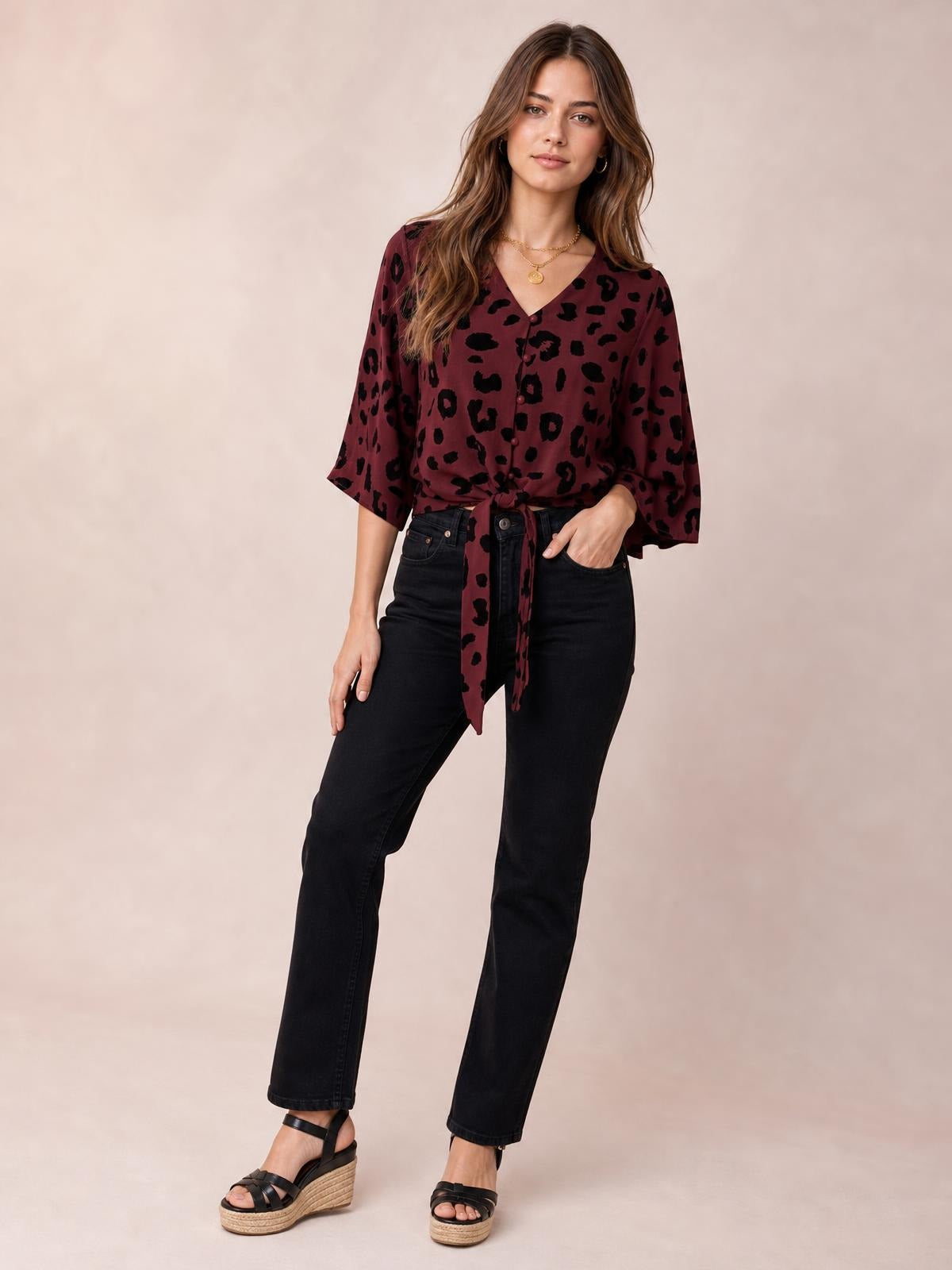 A model stands in a burgundy leopard-print V-neck blouse with a button front, three-quarter sleeves, and a tied waist creating a relaxed silhouette.